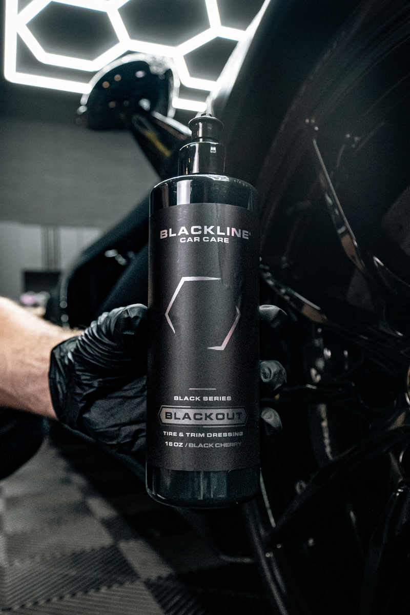 Black-gloved hand holding the Blackline Blackout trim and tire dressing bottle next to a vehicle wheel in a detailing bay.
