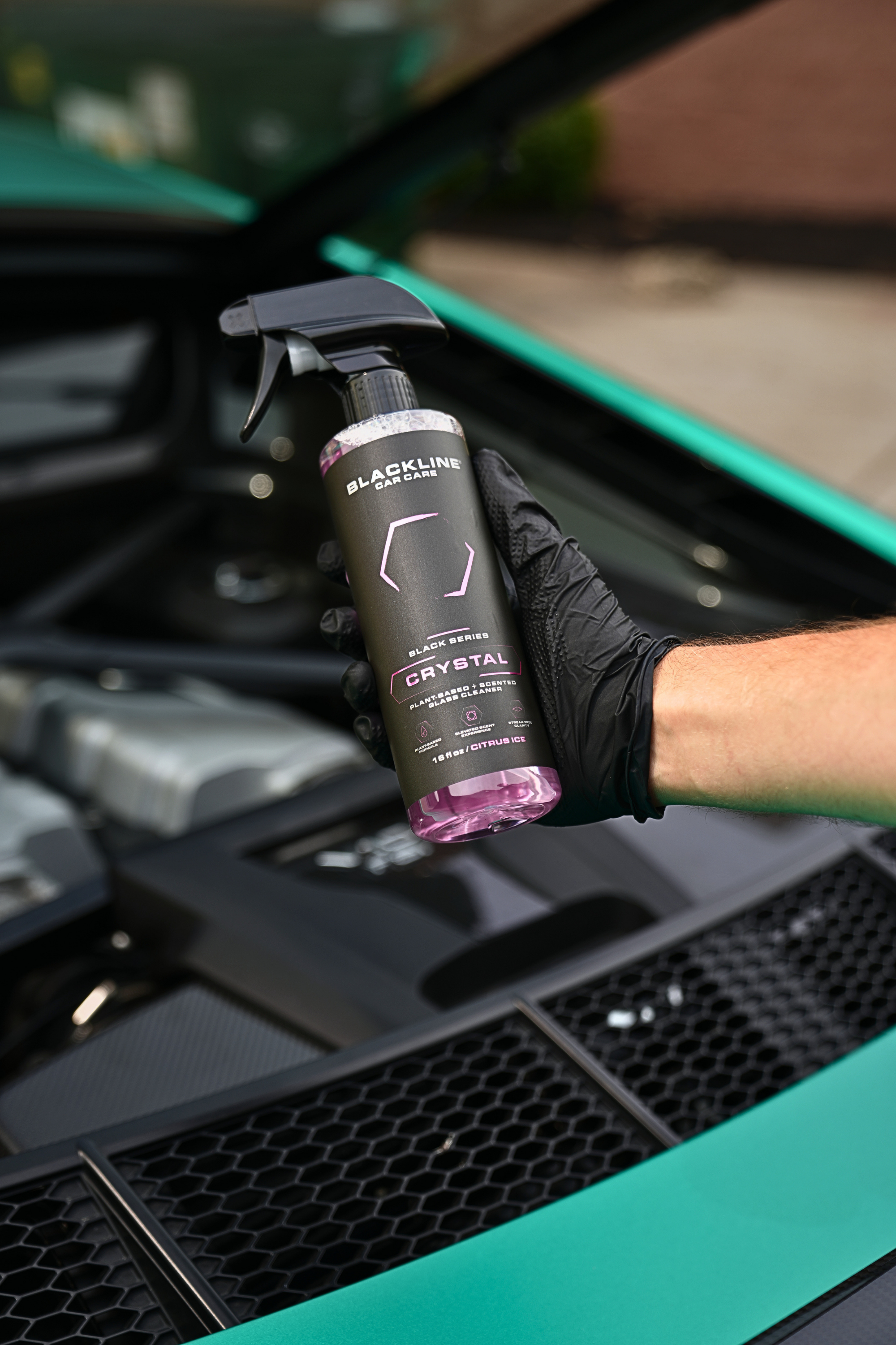 Hand holding Blackline Crystal Glass Cleaner bottle beside a clean car engine bay