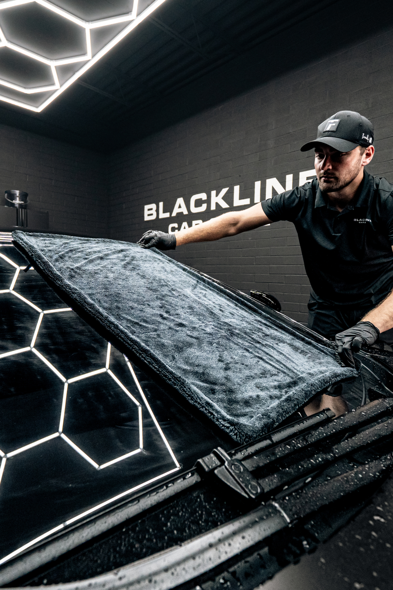 A detailer wiping water from a vehicle's windshield using the Blackline black twisted-loop microfiber drying towel inside a hexagon-lit detailing bay.